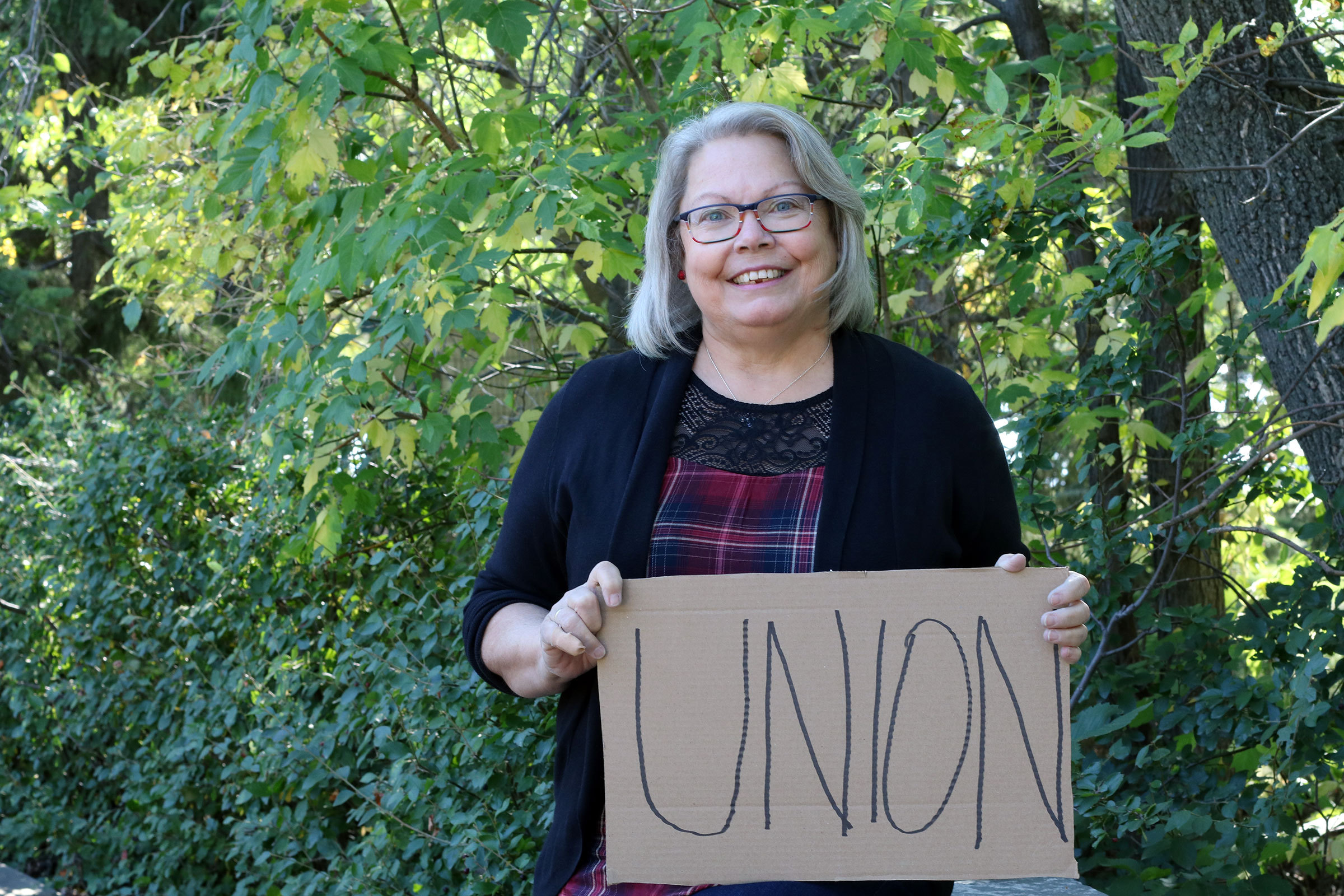 A woman with medium length grey hair and glasses smiling and holding a sign that reads UNION